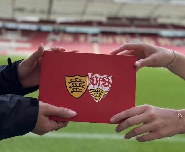 Zwei Hände halten gemeinsam einen roten Umschlag. Auf der Vorderseite sind das Wappen von Baden-Württemberg und das VfB Stuttgart Wappen abgebildet. Im Hintergrund das Stadion.