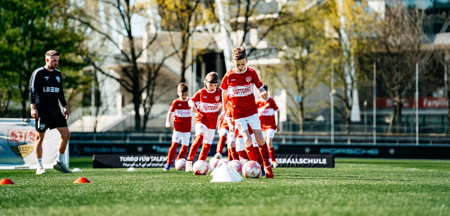 Mehrere Kinder im roten VfB Stuttgart Trikot trainieren mit Bällen. Daneben steht ein Trainer in schwarzer Trainingskleidung mit LBBW Logo