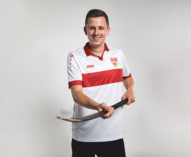 Ein Mann im VfB-Trikot balanciert einen Hockeyball auf dem Schläger – eine sportliche Kombination aus Fußball und Hockey.
