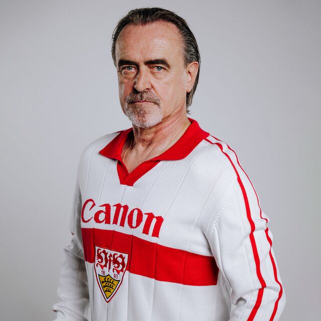 Günther Schäfer trägt das VfB Poloshirt Trikot Strick. Langarm Trikot in weiß mit rotem Brustring, rotem Polo-Kragen, großem VfB Wappen in der Mitte und Schriftzug Canon 