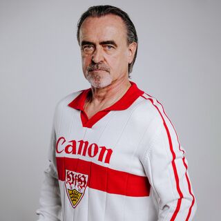 Günther Schäfer trägt das VfB Poloshirt Trikot Strick. Langarm Trikot in weiß mit rotem Brustring, rotem Polo-Kragen, großem VfB Wappen in der Mitte und Schriftzug Canon 