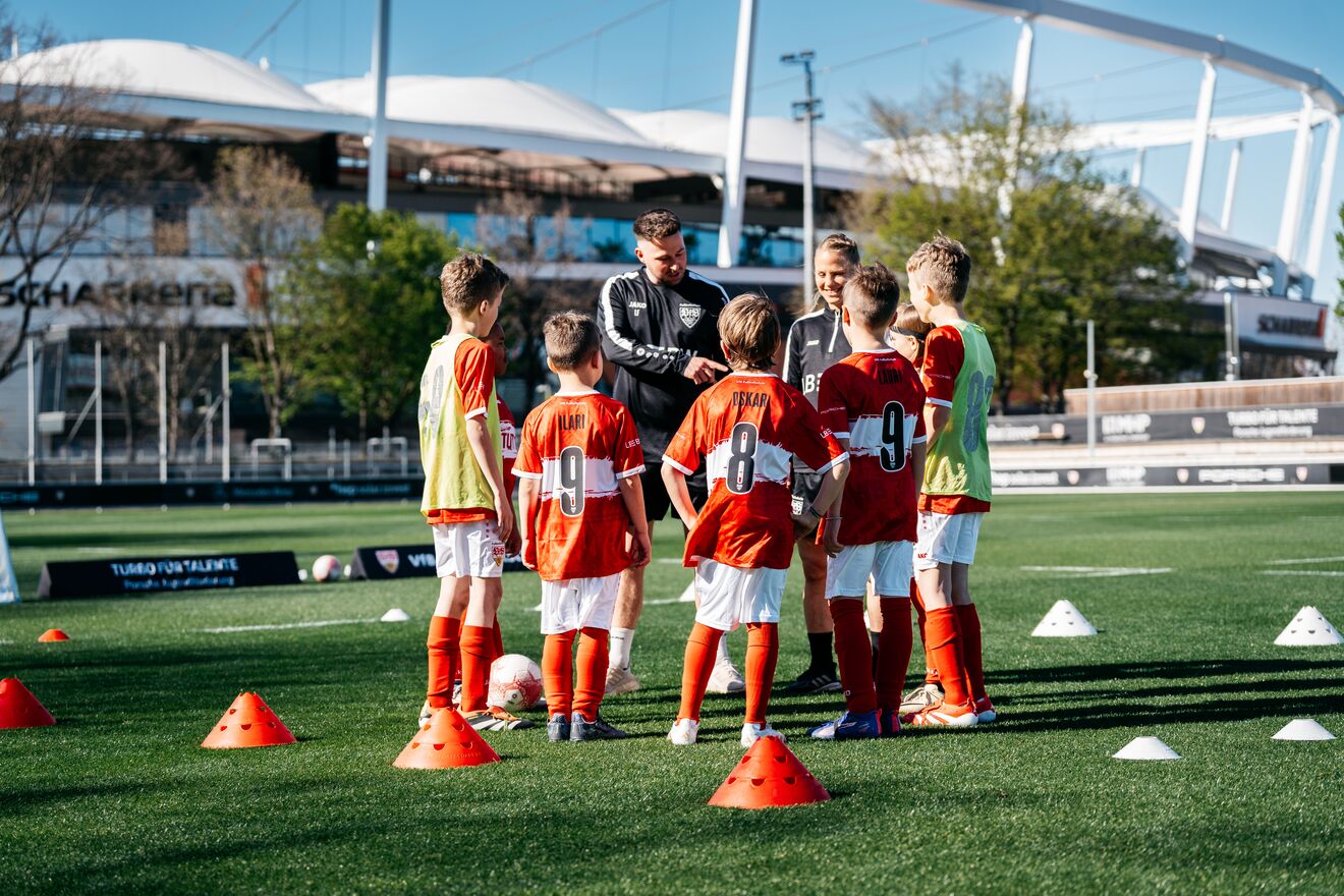 Mehrere Kinder im roten VfB Stuttgart Trikot stehen im Kreis um zwei Trainer in schwarzer Trainingskleidung. Im Hintergrund ist das Stadion zu sehen.