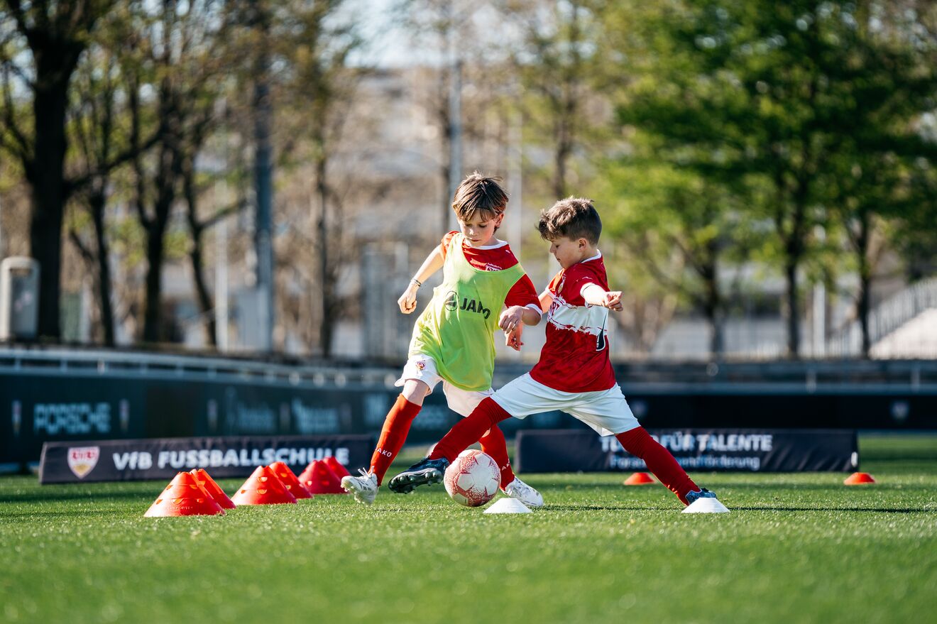 Zwei Kinder im roten VfB Stuttgart Trikot spielen im Training um den Ball. Eines trägt eine gelbe Trainingsweste. Ein Trainer in schwarzer Trainingskleidung mit LBBW Logo beobachtet die Szene.