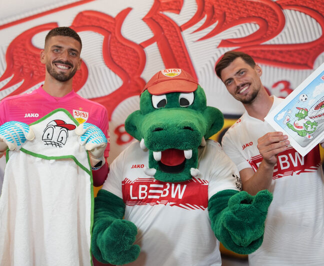 Das VfB-Maskottchen Fritzle steht zwischen zwei Spielern. Links wird ein Kinderhandtuch mit Fritzle-Motiv gezeigt, rechts ein Malbuch mit Stadion- und Maskottchenillustration.