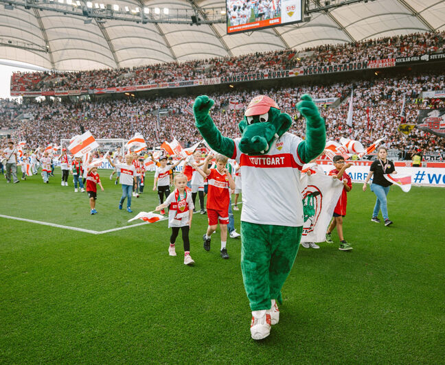 Das grüne Maskottchen Fritzle trägt rotes Cap und weißes VfB Trikot mit rotem Brustring. Er läuft mit erhobenen Armen über den Rasen, begleitet von Kindern mit VfB Fahnen. Im Hintergrund die volle Cannstatter Kurve.