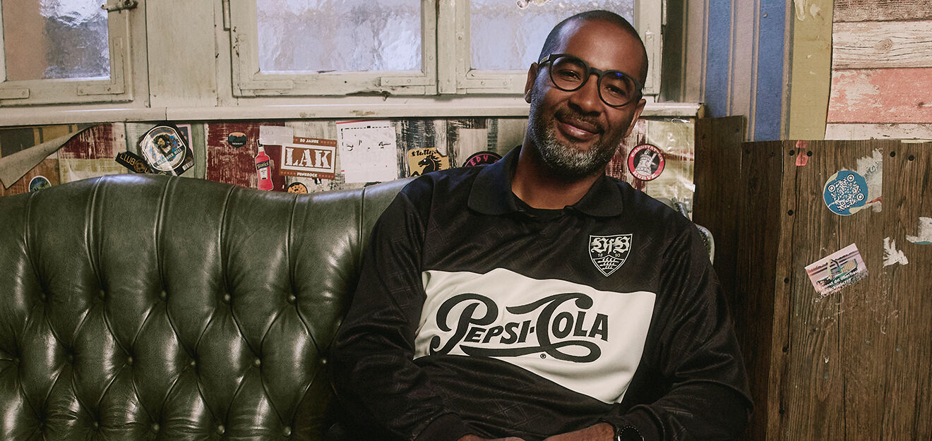 Cacau sitz auf einem Sofa und trägt das VfB Pepsi Trikot Retro langarm 