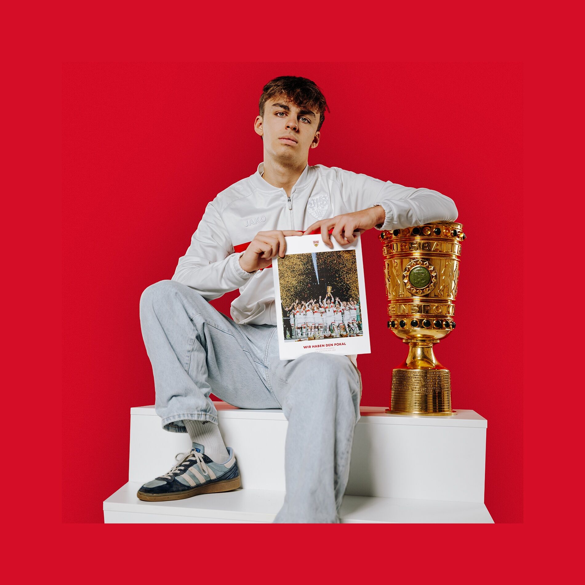 Junger Mann sitzt auf Stufen und hält den VfB Bildband Pokalsieger in seinen Händen. Neben ihm steht der DFB-Pokal. 