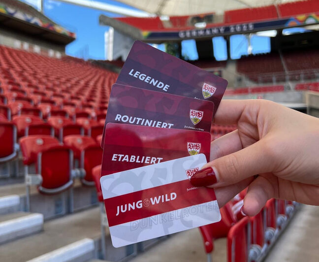 Eine Hand mit roten Fingernägeln hält vier VfB Stuttgart Mitgliedskarten vor leeren Tribünen. Die Karten tragen die Aufschriften ‚Legende‘, ‚Routiniert‘, ‚Etabliert‘ und ‚Jung & Wild‘.