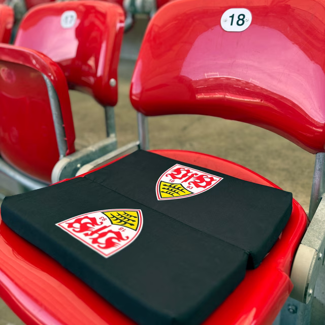 schwarzes Sitzkissen mit VfB Wappen liegt aufgeklappt auf einem roten Stadionsitz