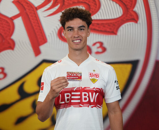 Ein Spieler im weißen VfB Stuttgart Trikot mit rotem Brustring hält eine rote VfB Jugendclub-Mitgliedskarte in die Kamera. Im Hintergrund ist vergrößert das VfB Wappen zu sehen.