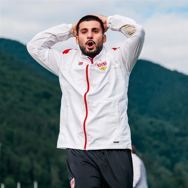 Ein Spieler trägt eine weiße VfB Trainingsjacke mit rotem Reißverschluss und Wappen auf der Brust. Er hat die Hände hinter dem Kopf verschränkt und blickt überrascht nach vorne.